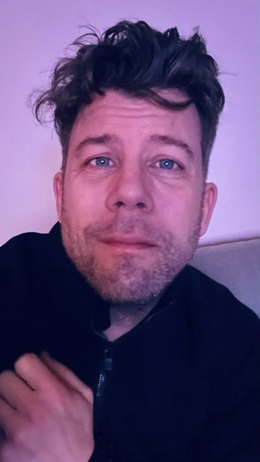 A close-up selfie of a man with dark, messy hair and blue eyes, wearing a black zip-up jacket, looking directly at the…