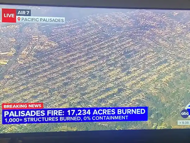 An aerial news broadcast shows a vast area of Pacific Palisades scorched by wildfire, with on-screen text stating 'PALISADES…