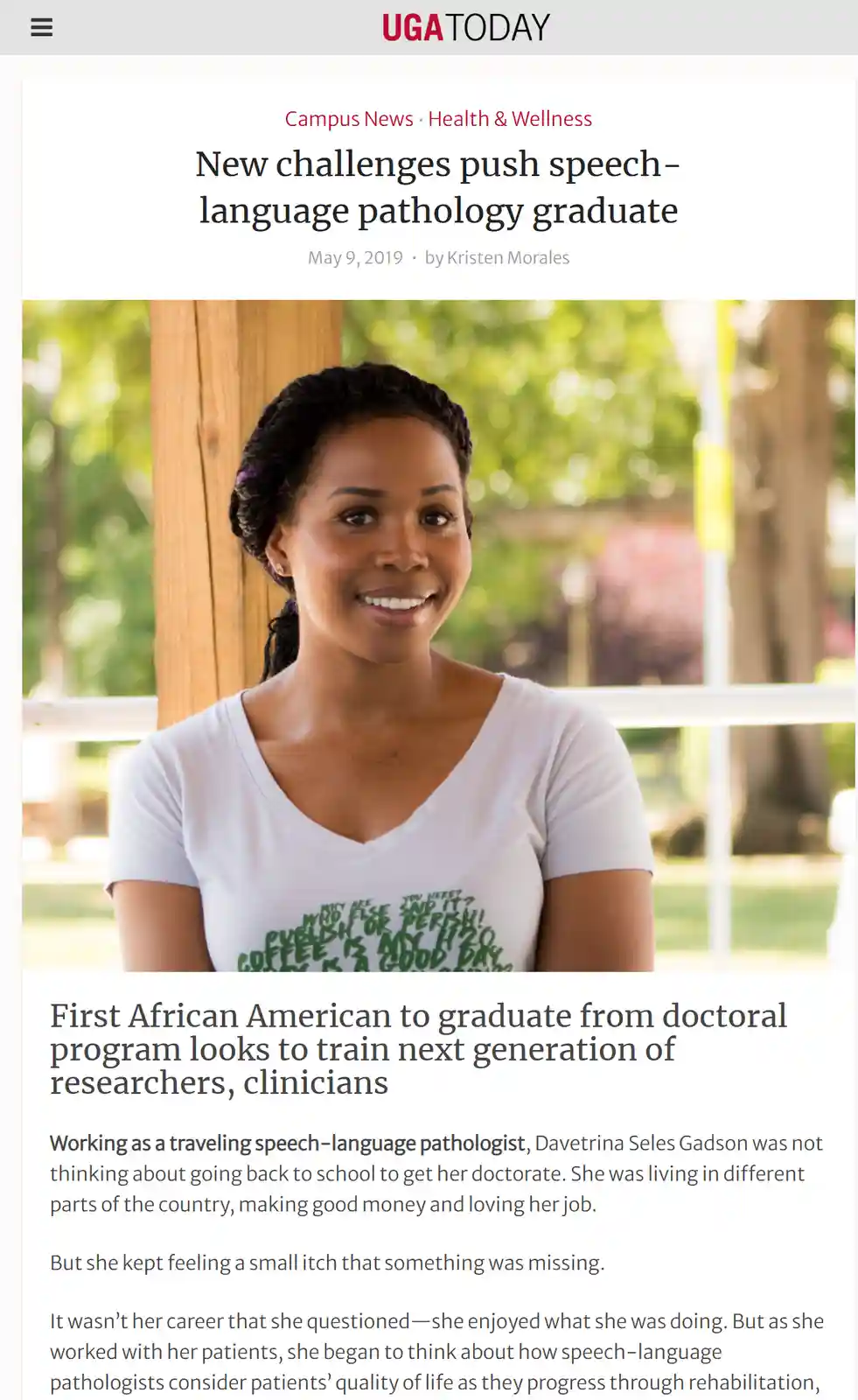 A smiling Black woman with braided hair wearing a white t-shirt with green text stands outdoors in front of a wooden…