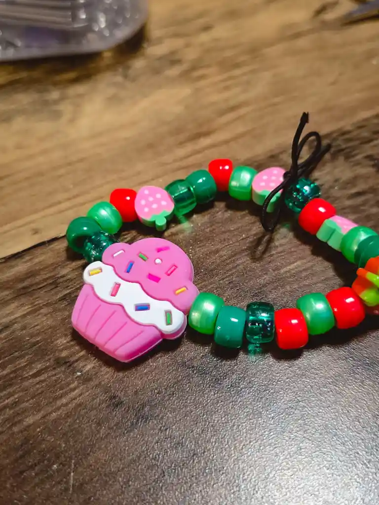 A close-up photo shows a colorful beaded bracelet on a wooden surface, featuring a large pink cupcake charm with white…