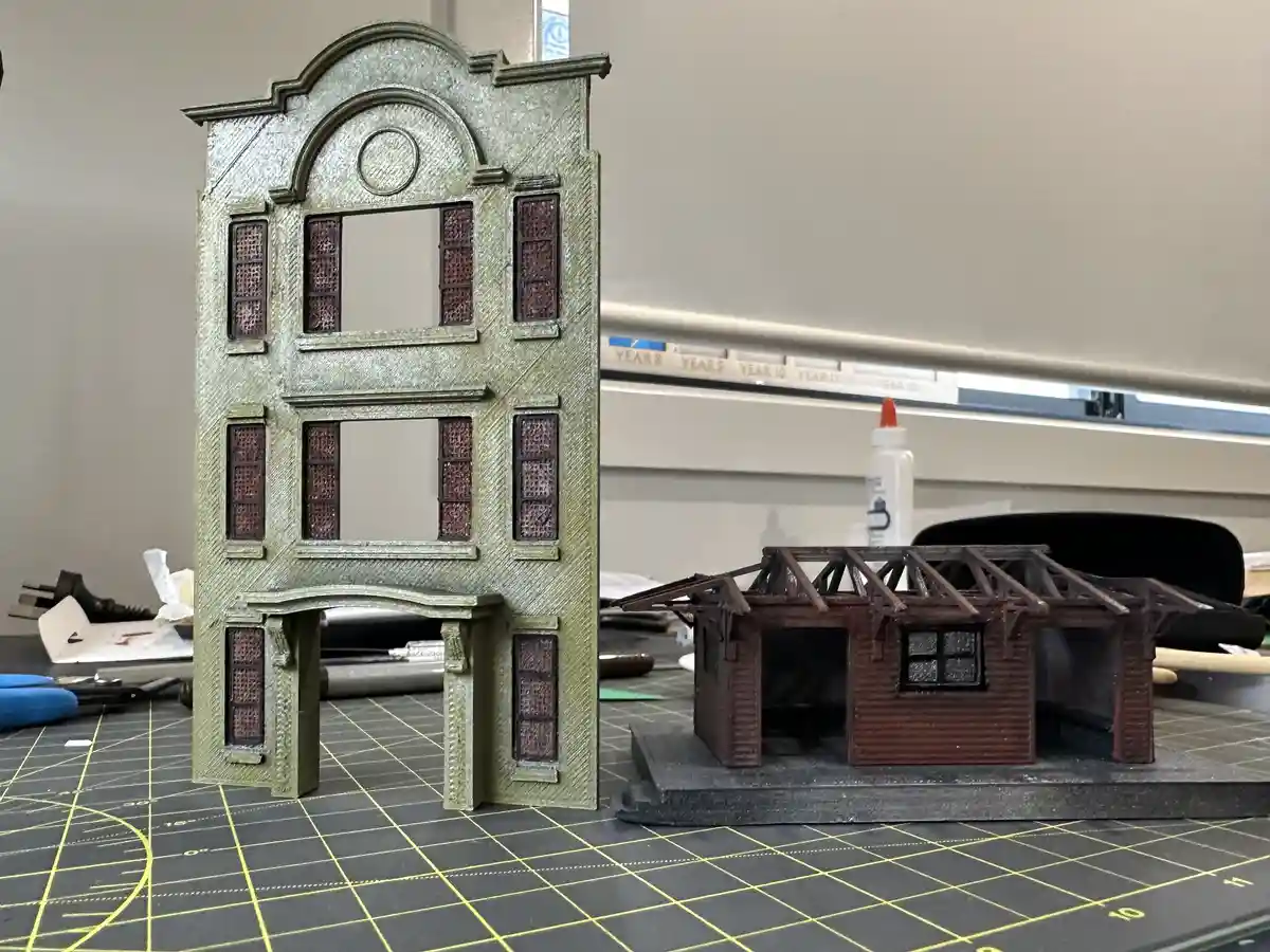 Two miniature buildings sit on a gridded crafting mat: a tall, ornate, fully detailed structure stands beside a smaller…