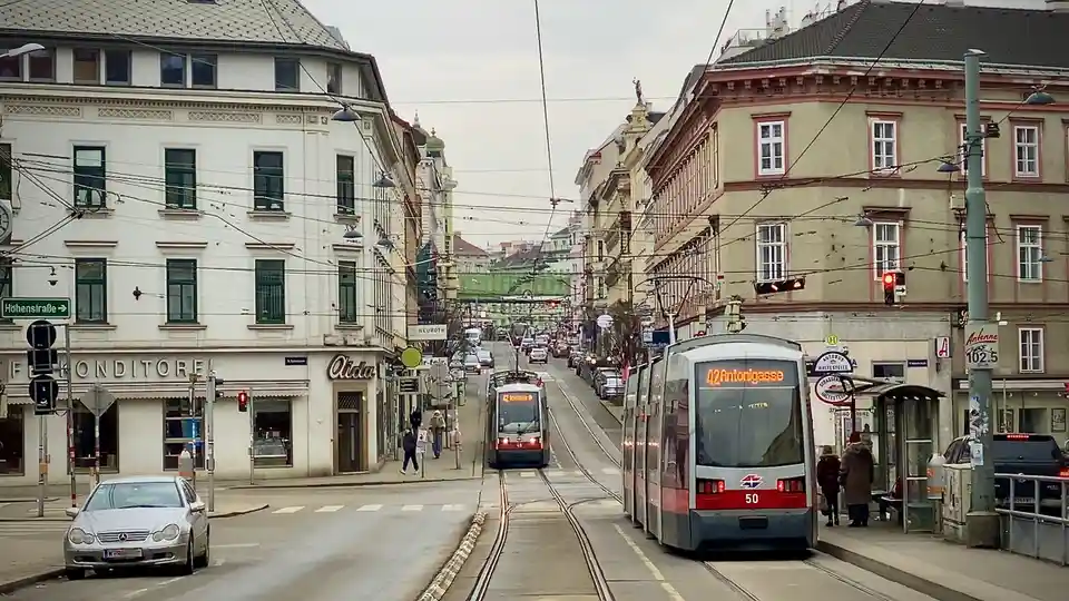 A street view of a European city with two trams on parallel tracks, one in the foreground and one further down the street…