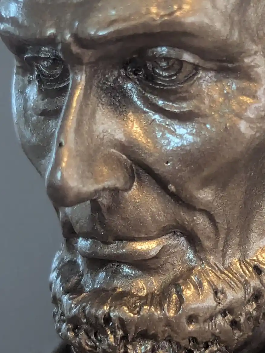 A close-up of a bronze statue's face, showing detailed wrinkles, a beard, and a stern expression. The overlaid text reads…