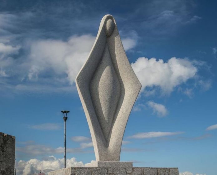A tall, abstract, grey stone statue with a stylized human-like form stands against a blue sky with white clouds. The statue…