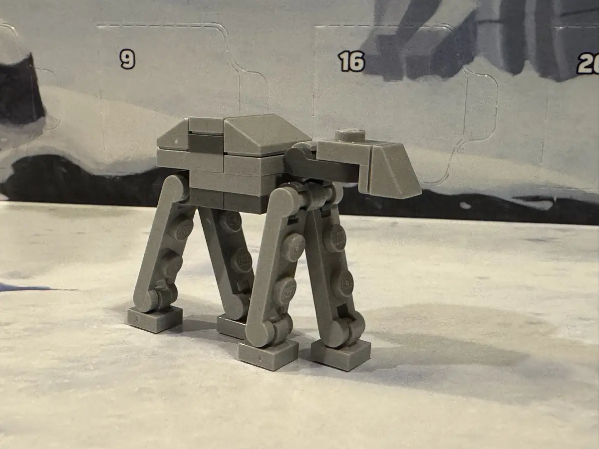 A small, gray LEGO model of an AT-AT walker stands on a snowy surface, with a printed LEGO box background showing numbers 9…