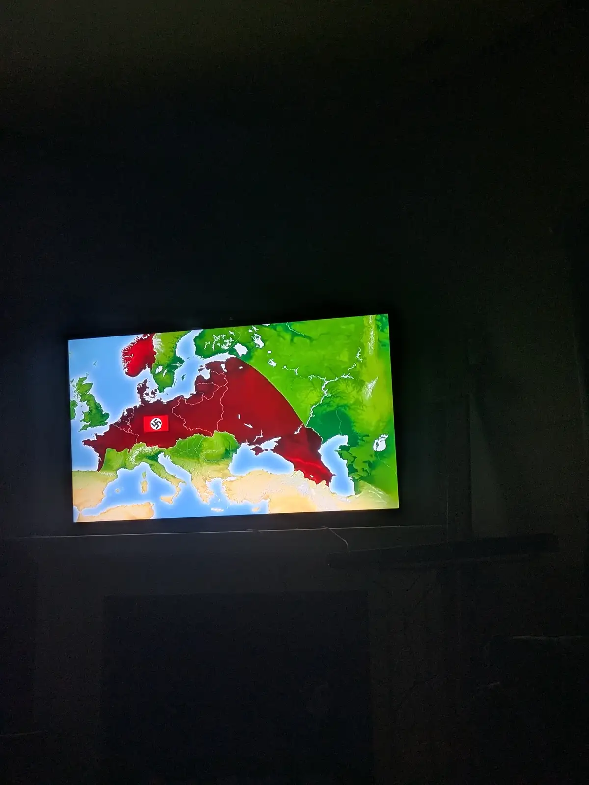 A dark room features a TV screen displaying a map of Europe with a large red area covering most of the continent, marked…