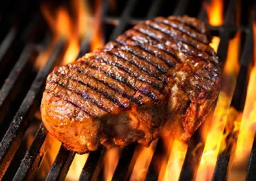 A close-up photo of a thick, juicy steak sizzling on a hot grill with visible flames licking around the meat, showing…