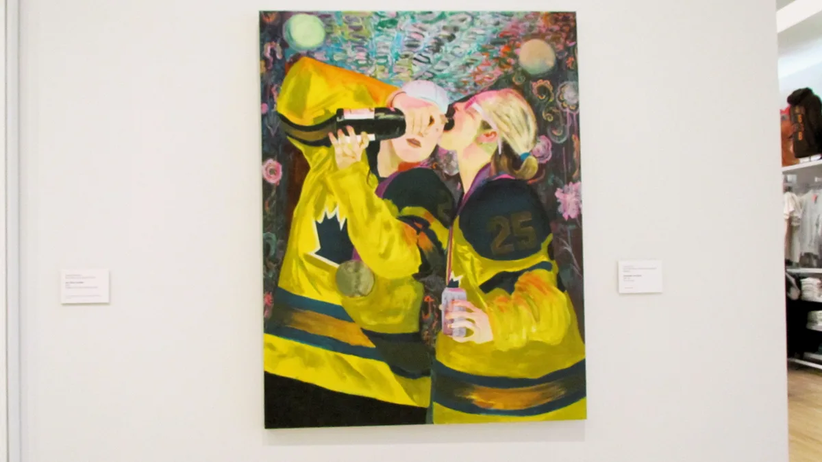 A painting of two people in yellow hockey jerseys drinking from a bottle, displayed in a gallery. The person on the left…