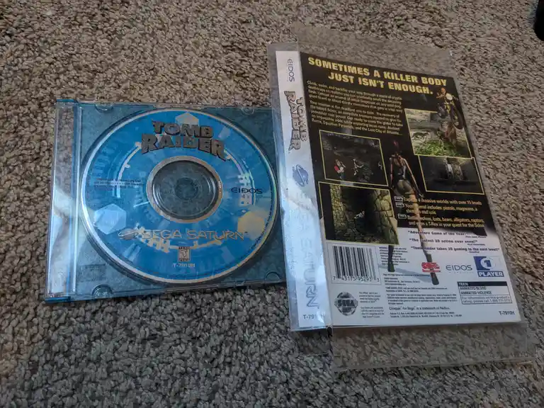 A physical copy of the Sega Saturn game 'Tomb Raider' is shown, with the game disc in its open case and the back cover…