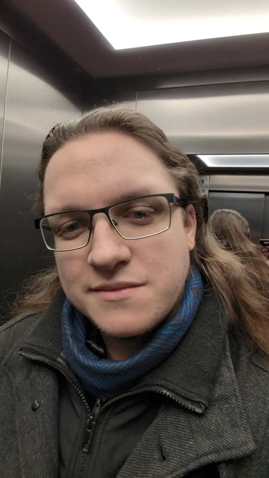 A person with long hair and glasses takes a selfie inside an elevator, wearing a dark coat and blue patterned scarf. The…