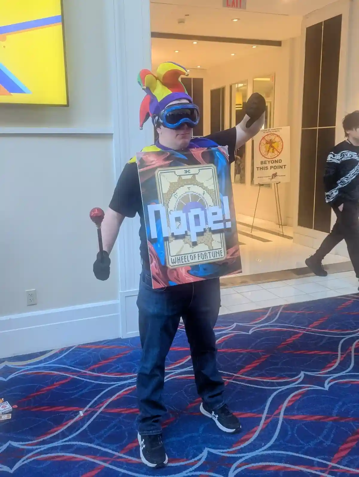 A person dressed as a jester with a colorful hat and large goggles holds a large sign that reads 'Nope! WHEEL OF FORTUNE'…