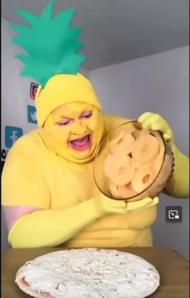 A person in a full-body yellow pineapple costume with a green spiky crown is holding a glass bowl of pineapple rings…