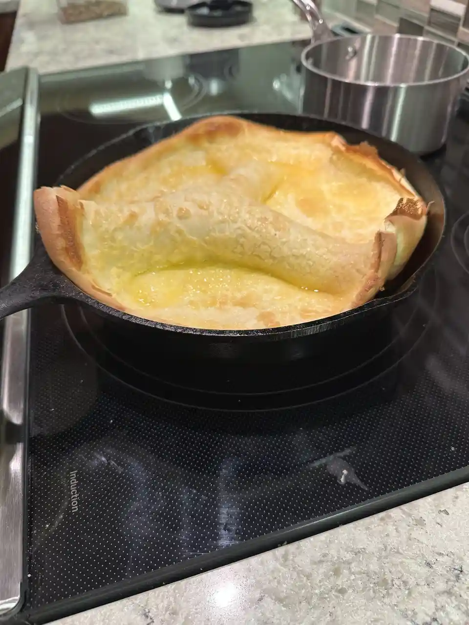 A golden-brown soufflé has collapsed inward in a black cast-iron skillet on an induction stovetop, with its once-raised…