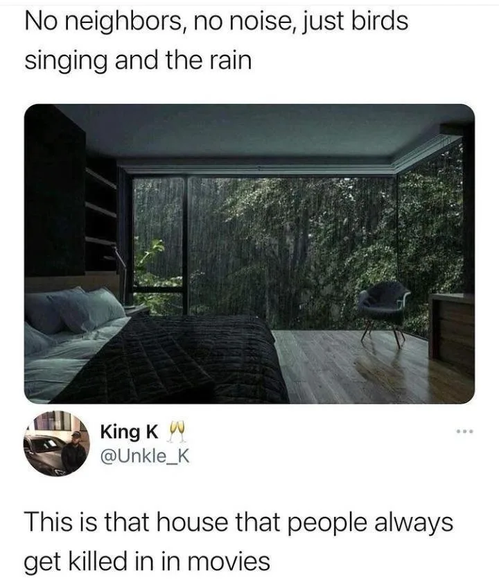 A dimly lit bedroom with a large glass wall overlooking a rainy forest. A bed with dark bedding is on the left, and a modern…