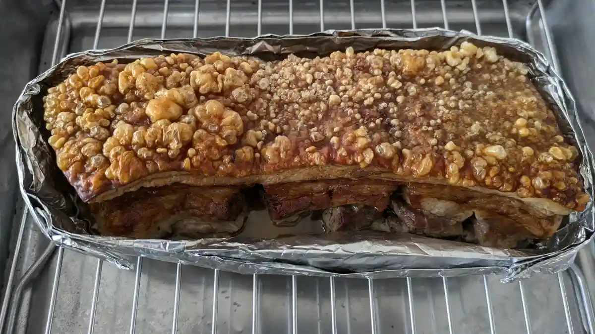 a large, roasted pork belly with a deeply caramelized, bubbly, crackling skin sits on aluminum foil inside a metal roasting…