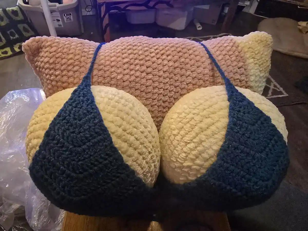 A handmade, chunky knit cat-shaped pillow with a peach body, cream-colored belly, and dark blue triangular straps resembling…