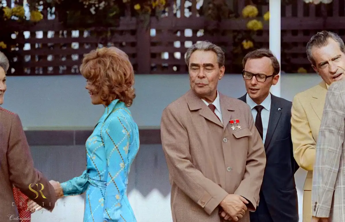 A colorized historical photo shows several men and women in formal attire at an outdoor event. A woman in a blue patterned…