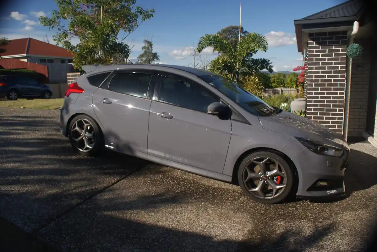 A gray Ford Focus ST with aftermarket wheels and a rear spoiler is parked on a gravel driveway in front of a house, covered…