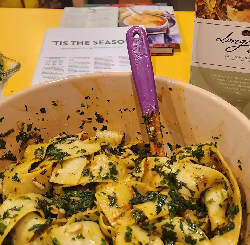 A close-up photo shows a large white bowl filled with wide pasta noodles mixed with chopped green herbs and pine nuts, with…