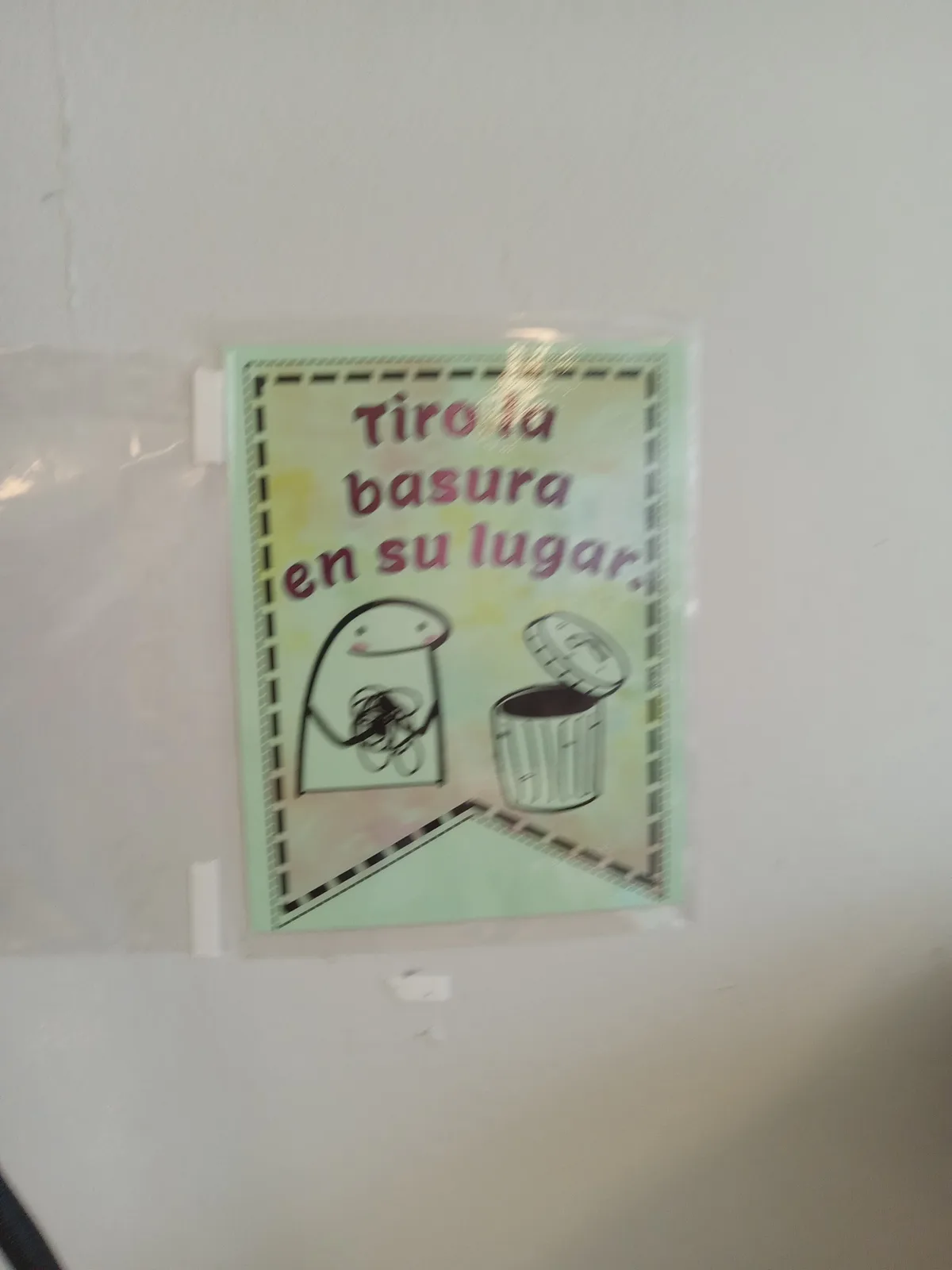 A light green poster taped to a white wall shows a cartoon character holding trash and walking toward an open trash can…