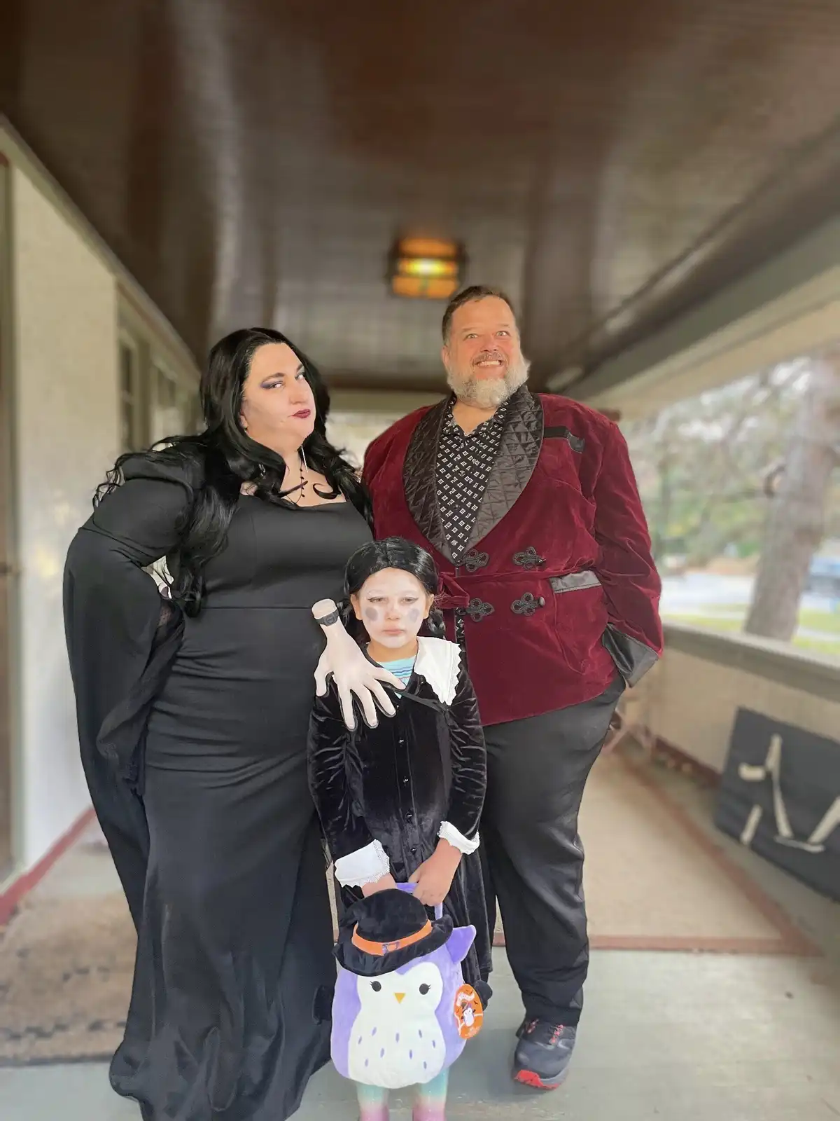 A family of three poses on a covered porch dressed in Halloween costumes. The woman on the left wears a black gothic gown…
