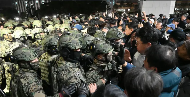a dense crowd of people surrounds a line of soldiers in full camouflage gear, helmets, and face coverings, with many…
