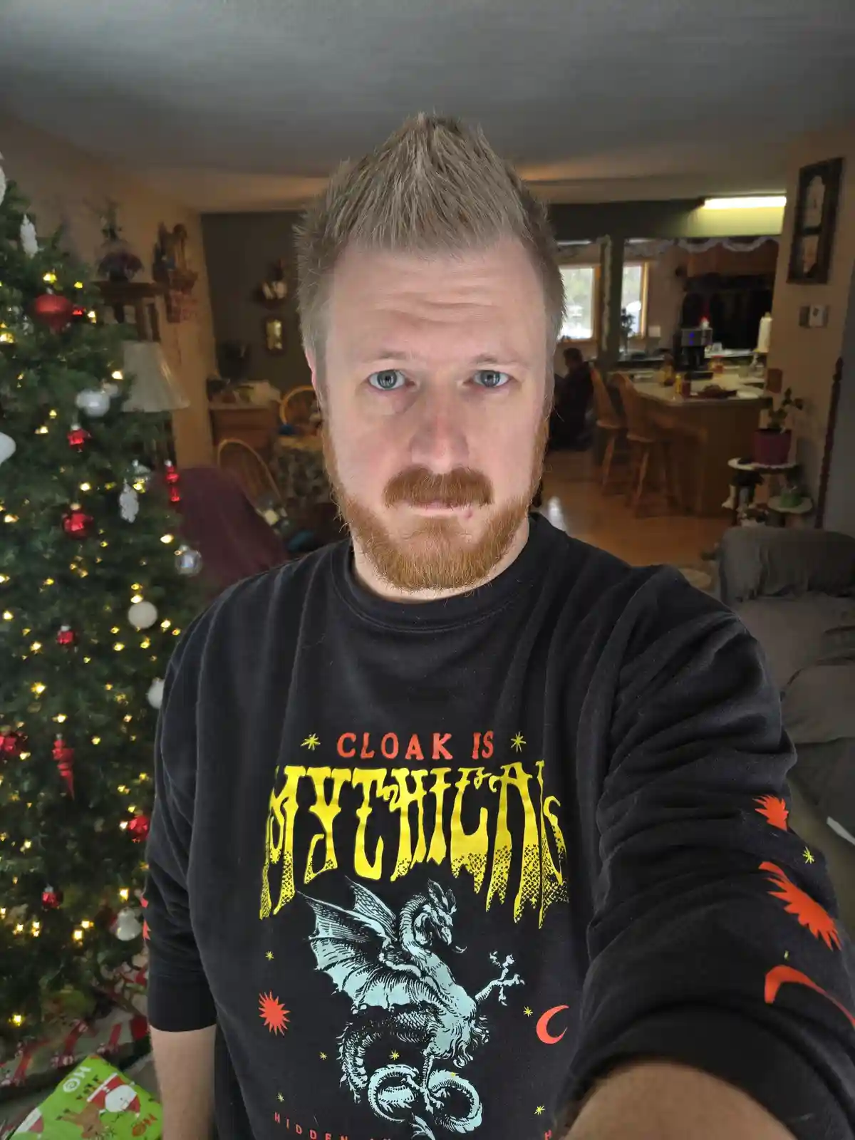 A man with a red beard and spiky blonde hair takes a selfie in front of a decorated Christmas tree, wearing a black…