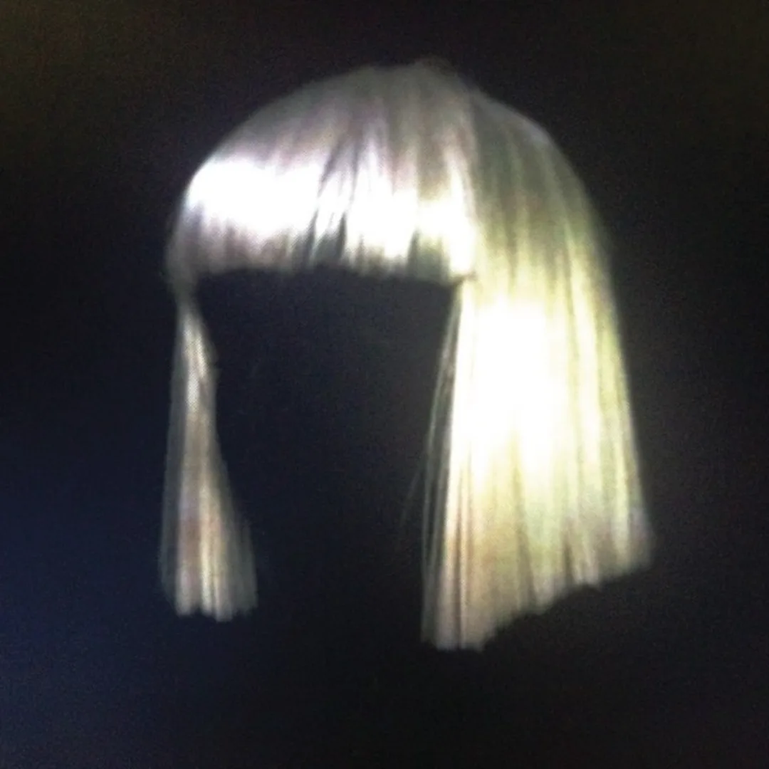 A blurry, low-resolution image shows a person wearing a bright, platinum blonde bob wig with bangs, set against a dark…