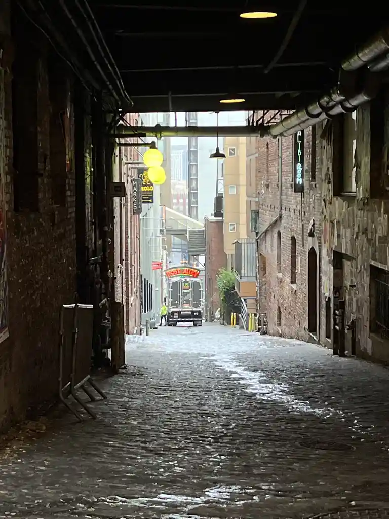A narrow, wet alleyway with brick walls and overhead pipes, leading to a truck with a red neon sign that reads 'MOST ALLEY'…