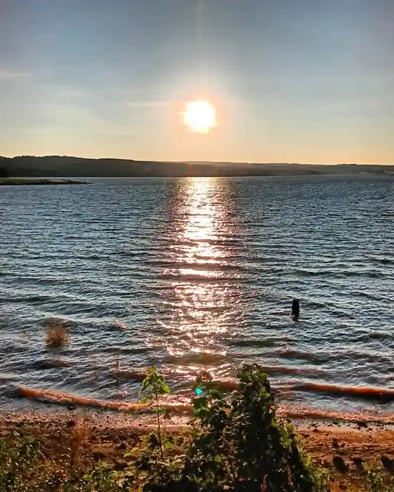 A serene sunset over a large body of water with the sun low on the horizon, casting a golden reflection across the rippling…