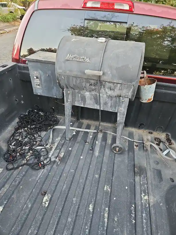 A heavily used Traeger pellet grill sits in the bed of a red pickup truck, surrounded by a tangled black power cord, a rusty…