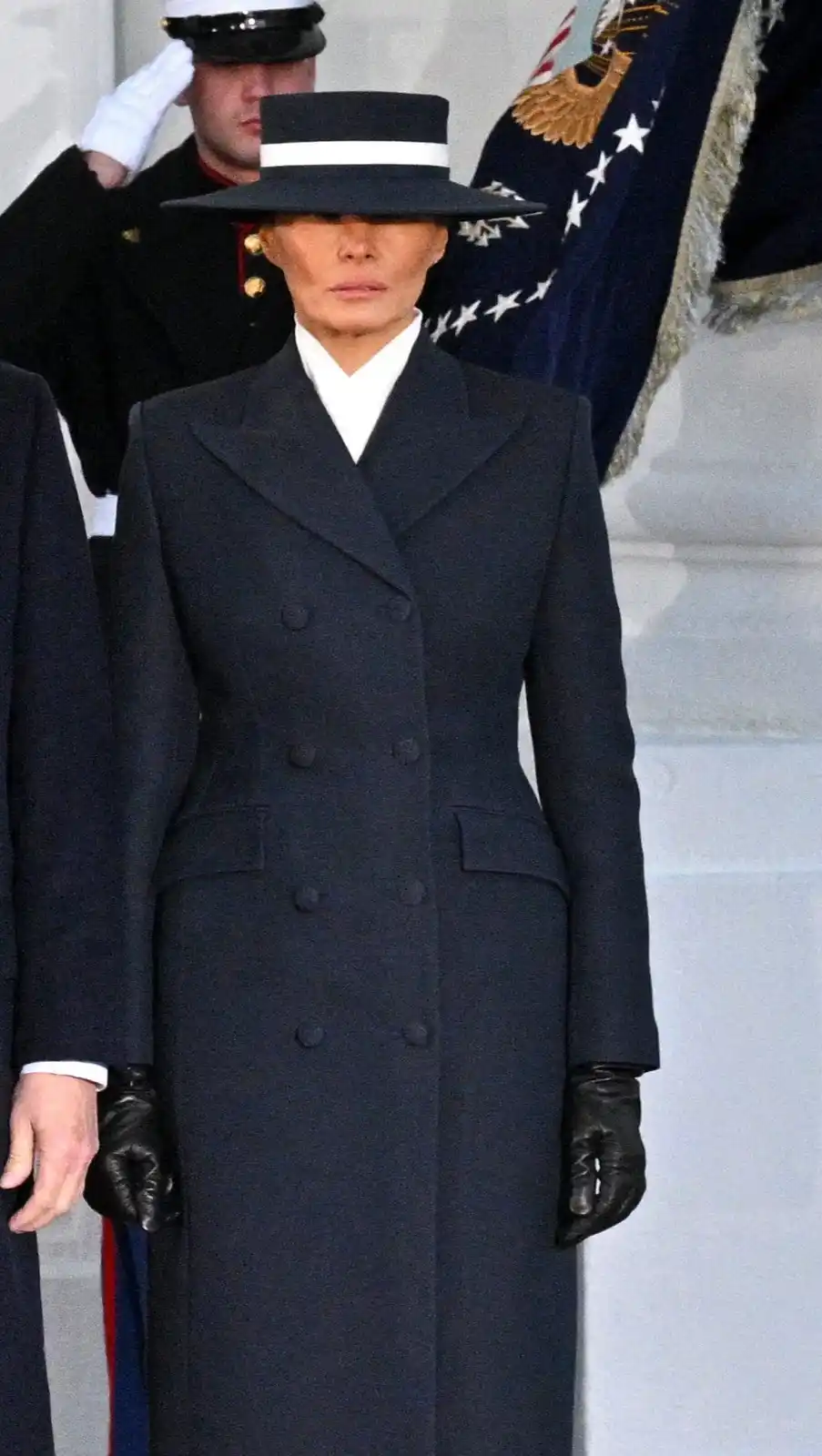 A woman in a dark navy double-breasted coat and black gloves stands solemnly, her face partially obscured by a wide-brimmed…