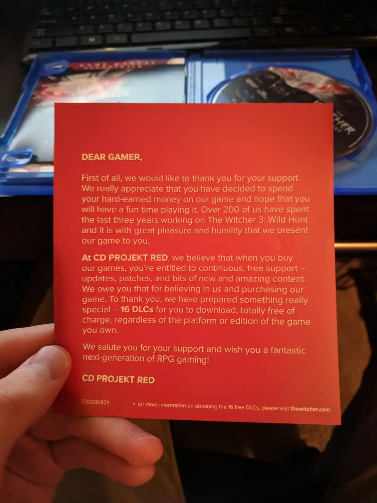 A hand holds a red card from CD Projekt Red thanking gamers for buying The Witcher 3: Wild Hunt, offering 16 free DLCs as a…