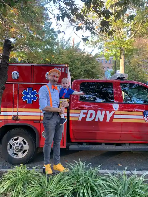 A man in bright orange suspenders, a bowtie, and glasses holds a toddler in front of a red FDNY emergency vehicle. The…