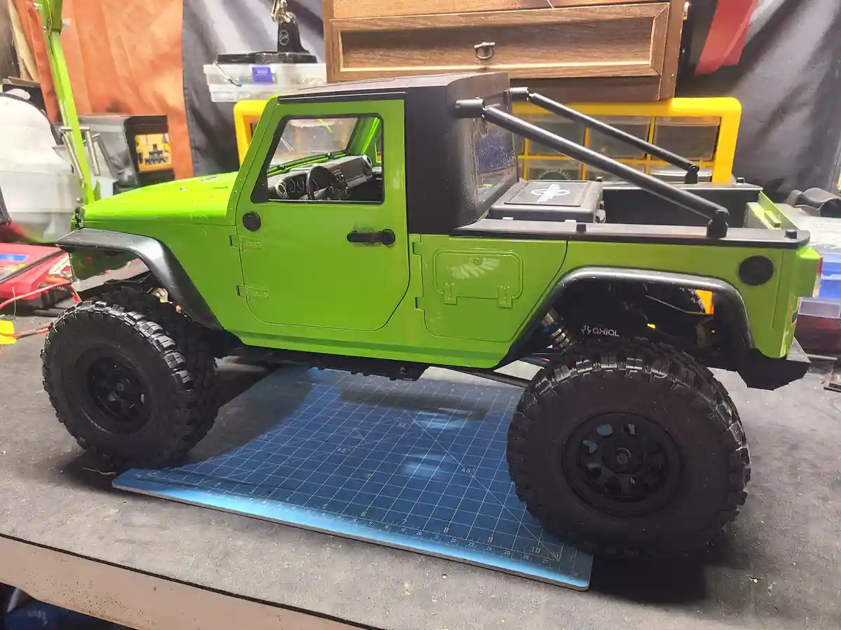 A bright green remote-controlled Jeep model with black tires and roll bars sits on a blue cutting mat in a workshop. The…