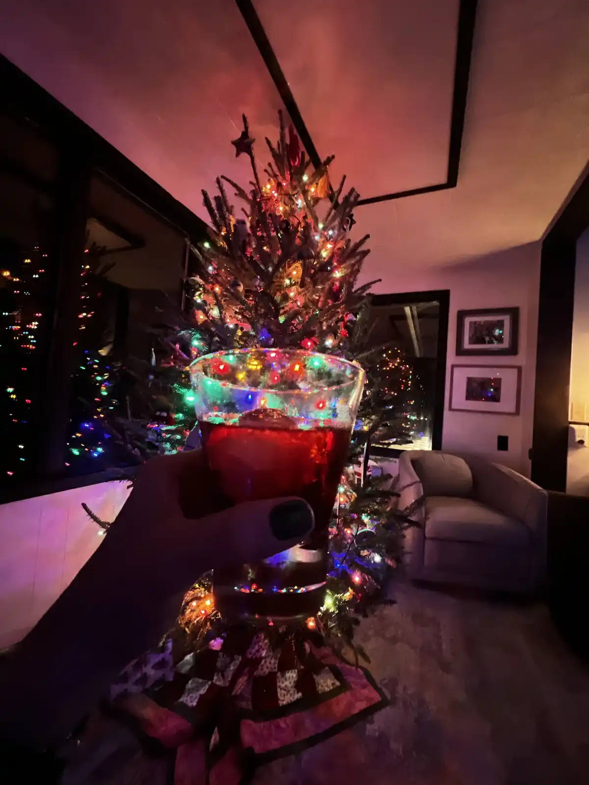 A hand holds a glass of red drink in front of a decorated christmas tree with colorful lights, in a cozy living room with a…