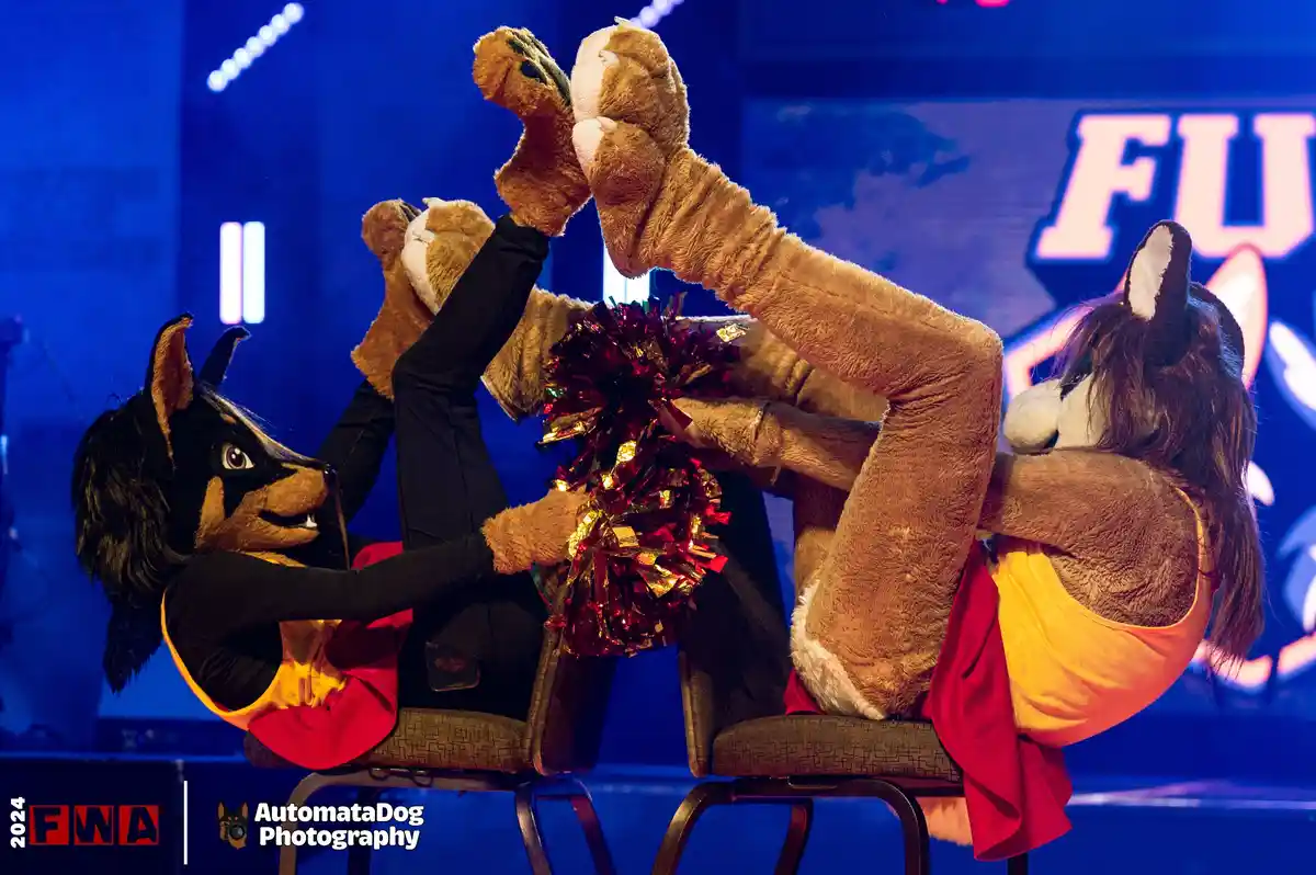 Two people in furry animal costumes—one resembling a black and tan dog, the other a brown fox—are seated on chairs…