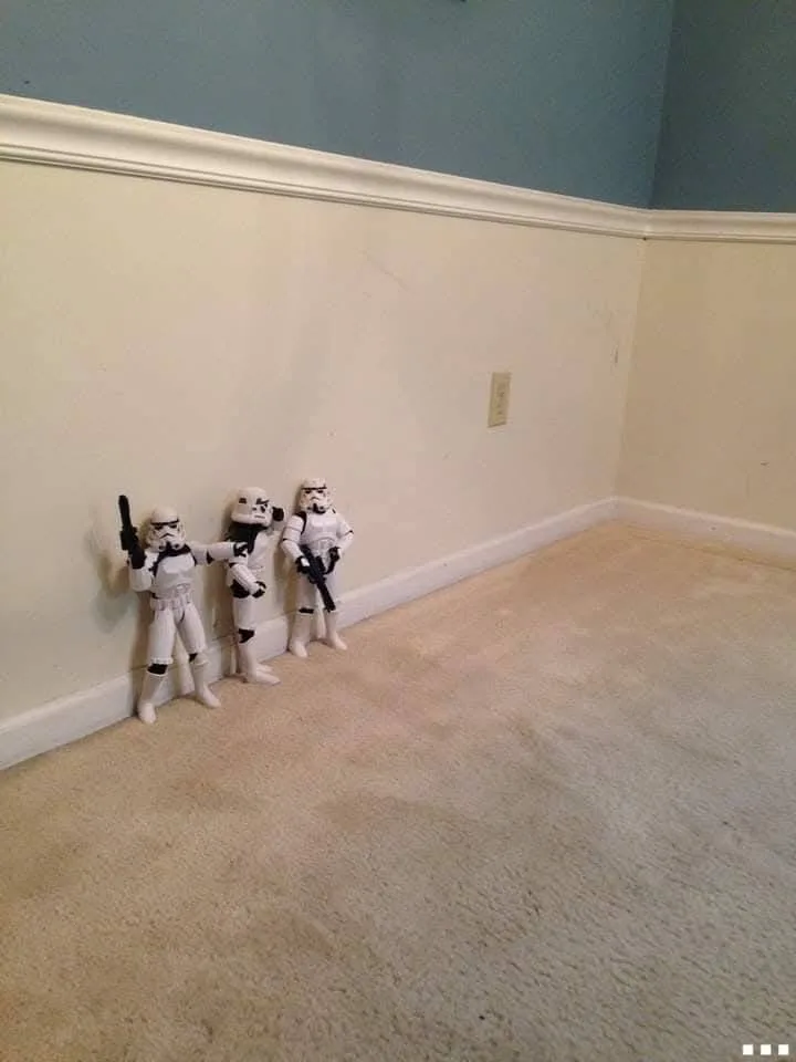 Three Star Wars Stormtrooper action figures stand in a corner of a carpeted room, leaning against the wall with one figure…