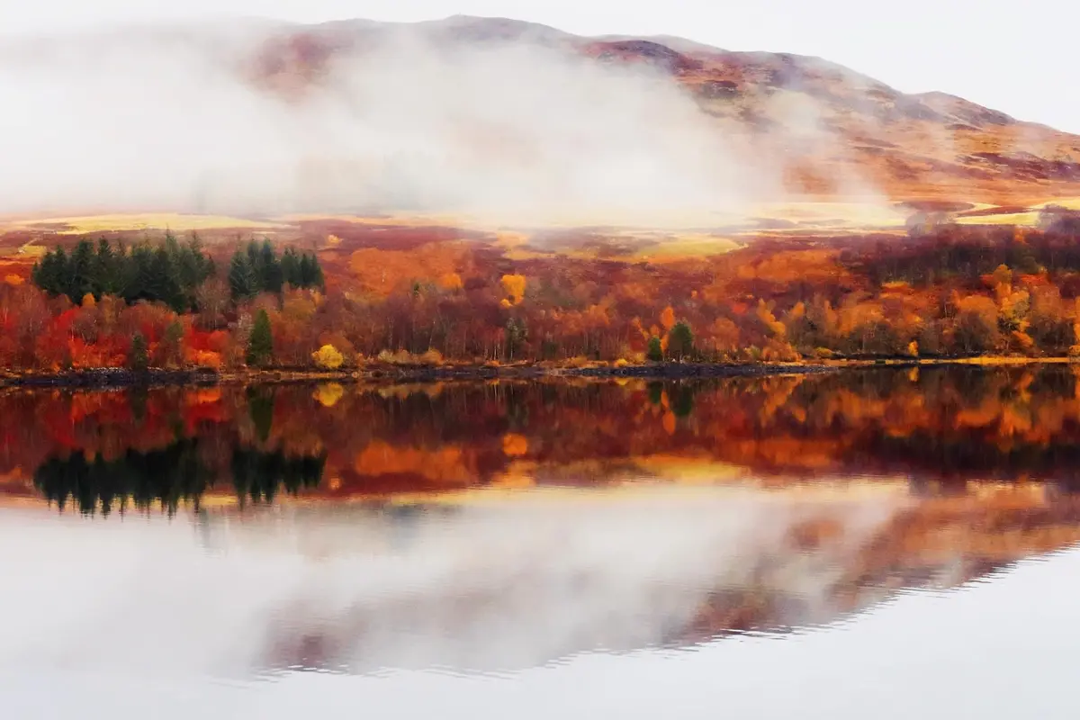A serene autumn landscape shows a calm lake reflecting vibrant orange, red, and yellow foliage on the shore, with misty…