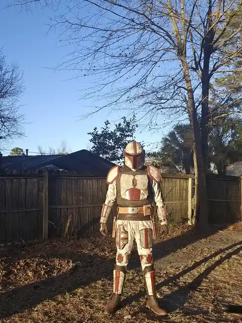 A person in a weathered, white-and-red Mandalorian armor costume stands in a backyard with a wooden fence and bare trees…