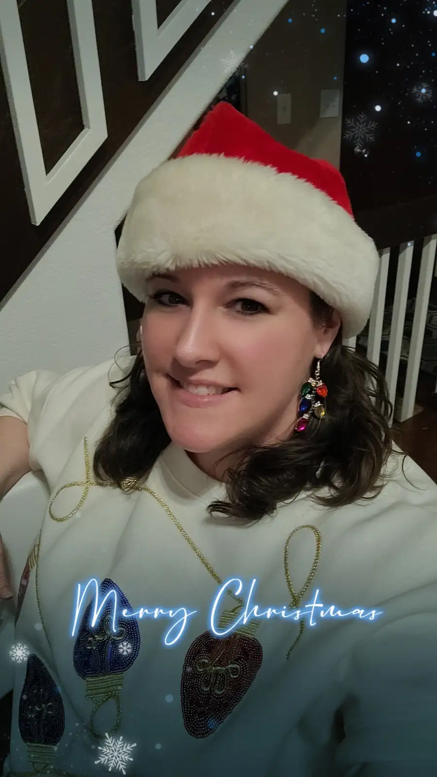 A woman with dark hair smiles while wearing a red and white Santa hat and a light-colored holiday sweater decorated with…