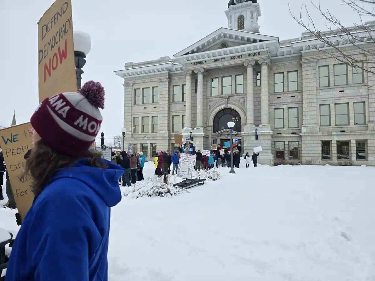 A snowy protest scene outside the Missoula County Courthouse, where demonstrators hold signs like 'Defend Democracy Now' and…