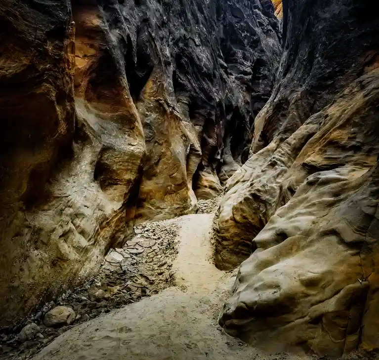 A narrow, winding canyon path with towering, textured rock walls in earthy tones of brown and gray, leading into the…
