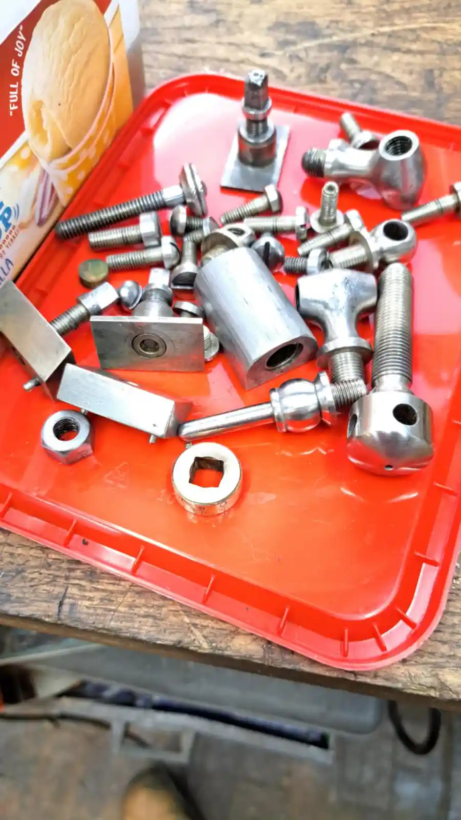 A red plastic tray holds a jumble of assorted metal hardware like bolts, nuts, and fittings, with a vanilla ice cream…