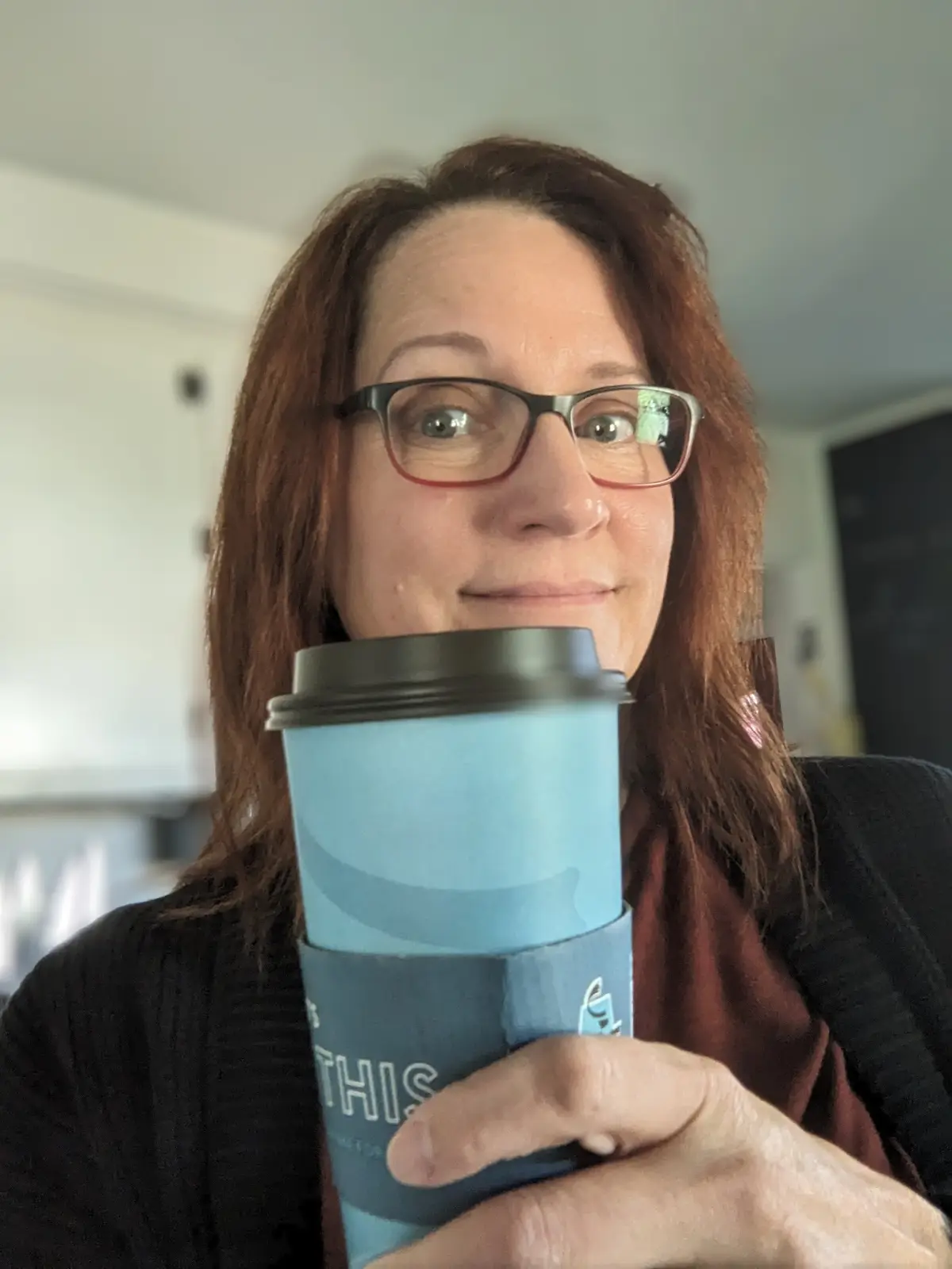 A woman with glasses holds a blue coffee cup in front of her face, obscuring her mouth and nose, while her eyes look…