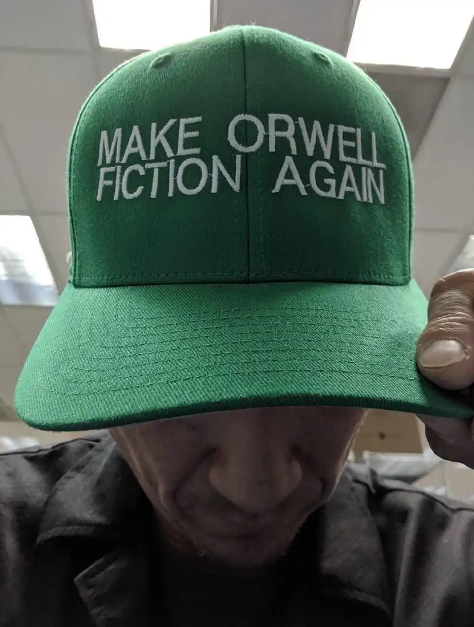 A close-up photo shows a person holding up a green baseball cap with white embroidered text that reads 'MAKE ORWELL FICTION…