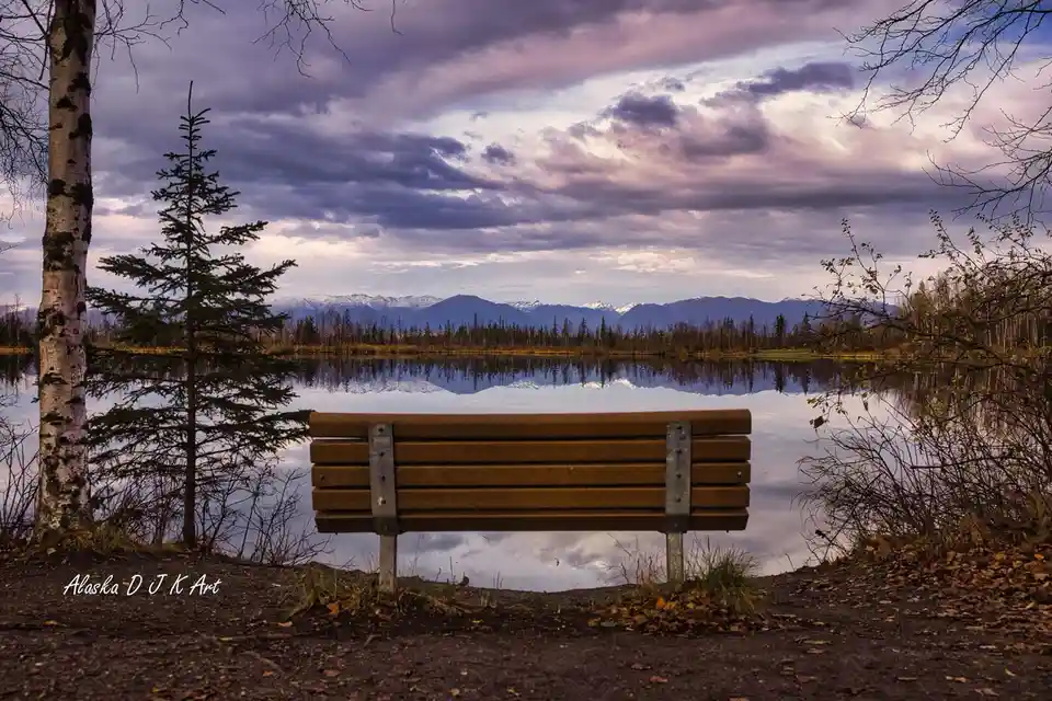 A wooden park bench faces a calm lake that perfectly mirrors distant snow-capped mountains under a dramatic purple and grey…