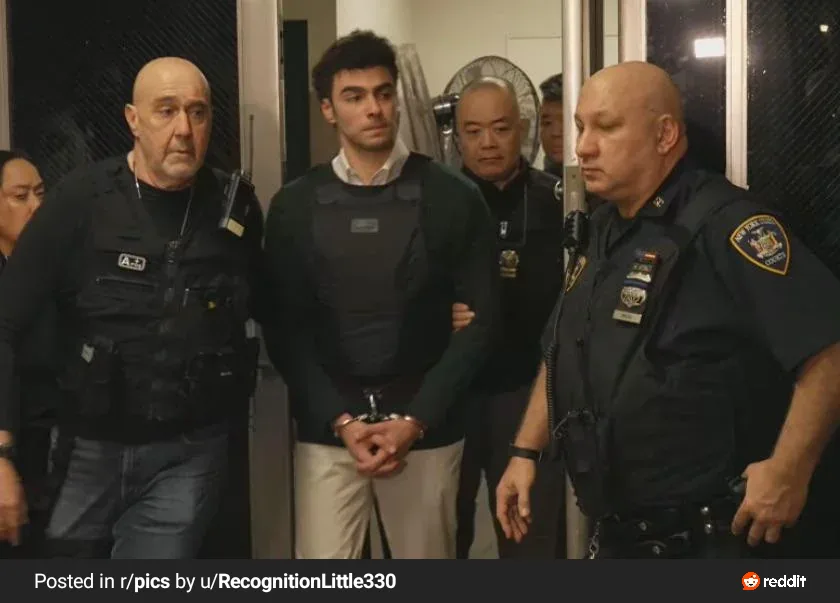 A man in a dark vest is flanked by two police officers, his hands cuffed in front of him. The overlaid text reads 'i'm not a…
