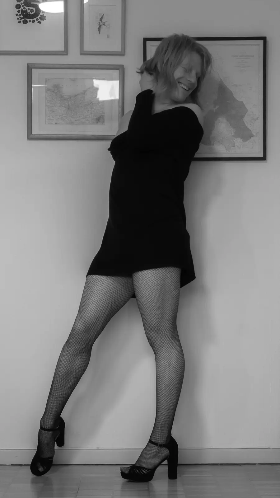 A black-and-white photo of a woman in a black dress, fishnet stockings, and heels, leaning against a wall with framed maps…