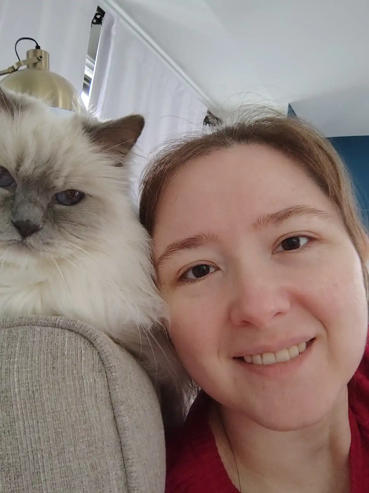A selfie shows a smiling woman with a fluffy, long-haired cat with blue eyes resting on her shoulder. The cat has a neutral…